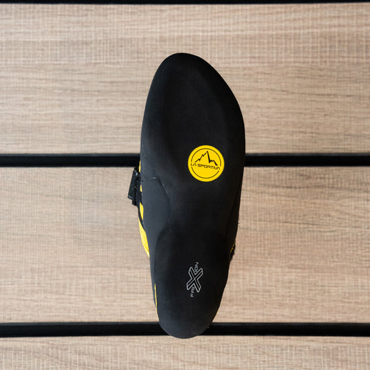 The sole of a climbing shoe, La Sportiva Tarantula Junior, hanging on a black hook against a textured metallic background.