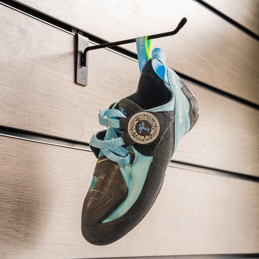 A climbing shoe, Tenaya Indalo hanging on a black hook against a textured metallic background.