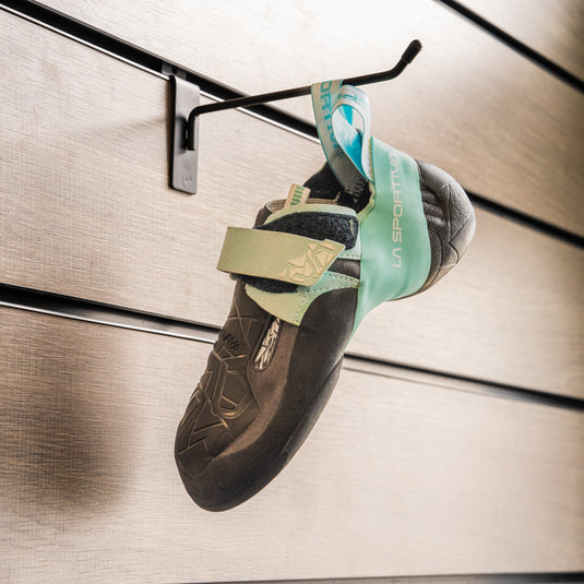 A climbing shoe, La Sportiva Skwama womens vegan hanging on a black hook against a textured metallic background.