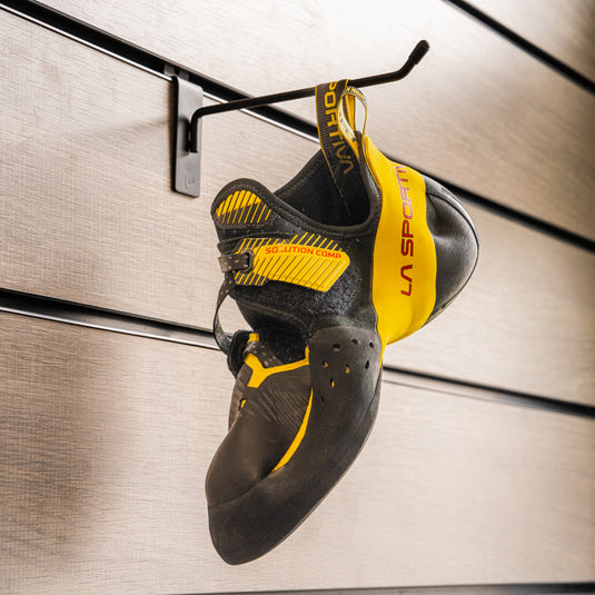 A climbing shoe, La Sportiva Solution comp, hanging on a black hook against a textured metallic background.