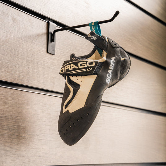 A climbing shoe, Scarpa Drago LV, in white and black design hanging on a black hook against a textured metallic background.