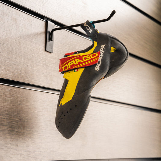 A climbing shoe, Scarpa Drago, in yellow and red design, hanging on a black hook against a textured metallic background.