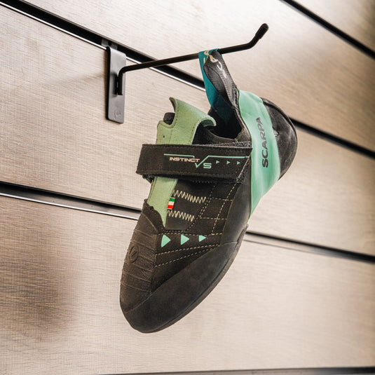 A climbing shoe, Scarpa Instinct VS WMN, hanging on a black hook against a textured metallic background.