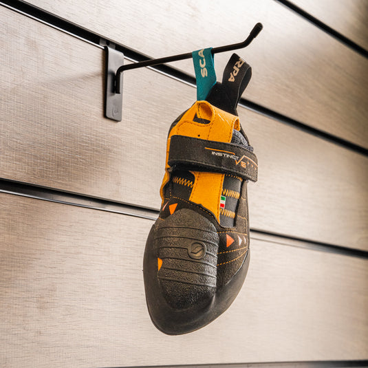 A climbing shoe, Instinct VS, hanging on a black hook against a textured metallic background.