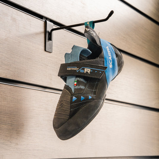 A climbing shoe, Scarpa Instinct VSR, hanging on a black hook against a textured metallic background.