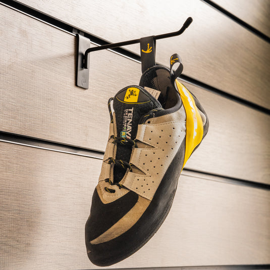 A climbing shoe, Tenaya Tarifa hanging on a black hook against a textured metallic background.