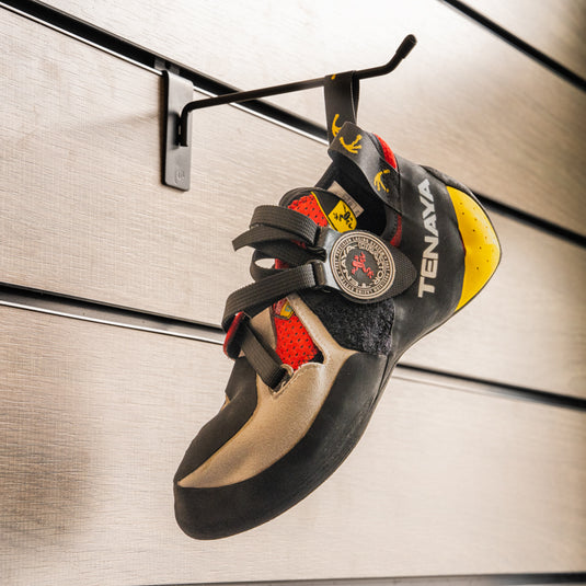 A climbing shoe, Tenaya Iati hanging on a black hook against a textured metallic background.