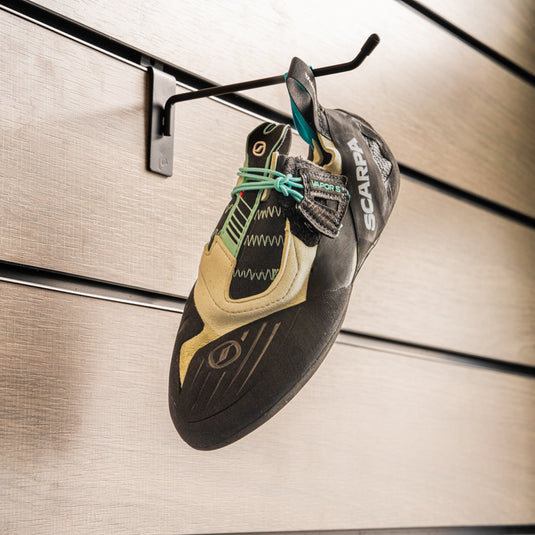 A climbing shoe, Scarpa Vapour S Women, hanging on a black hook against a textured metallic background.