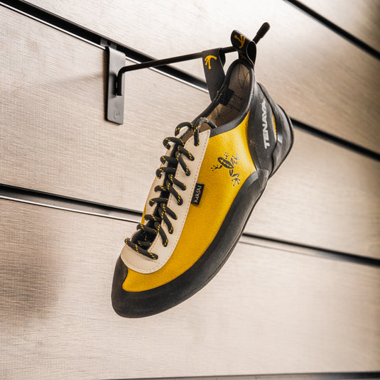 A climbing shoe, Tenaya Masai hanging on a black hook against a textured metallic background.