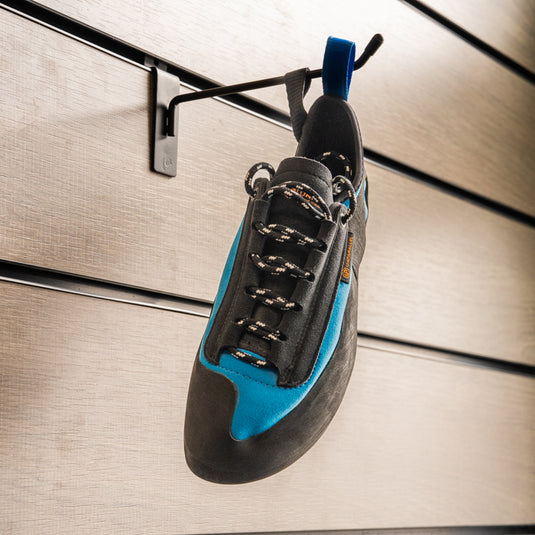 A climbing shoe, Unparallel Up Lace hanging on a black hook against a textured metallic background.