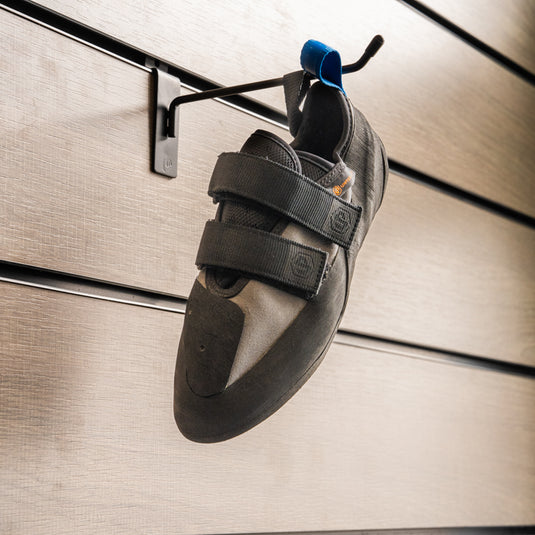 A climbing shoe, Unparallel Up Rise hanging on a black hook against a textured metallic background.