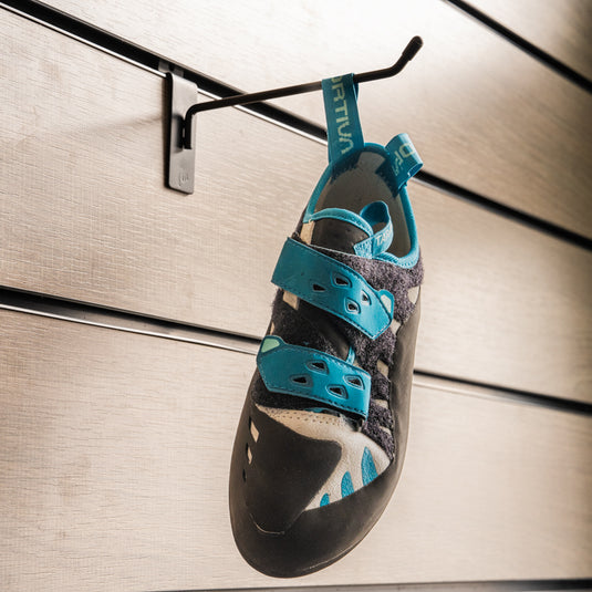 A climbing shoe, La Sportiva Tarantula Boulder womens, hanging on a black hook against a textured metallic background.