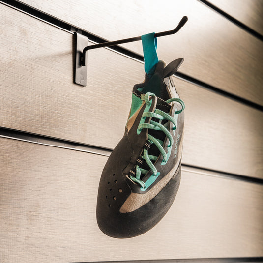 A climbing shoe, Scarpa Veloce Lace wmn hanging on a black hook against a textured metallic background.