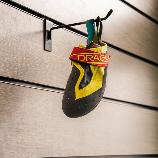 A climbing shoe, Scarpa Drago Kids hanging on a black hook against a textured metallic background.