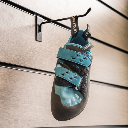 A climbing shoe, La Sportiva Tarantula velcro womens hanging on a black hook against a textured metallic background.