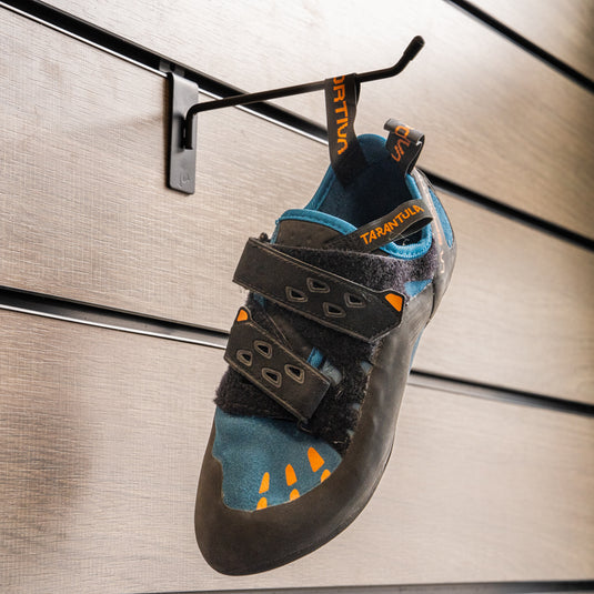A climbing shoe, La Sportiva Tarantula velcro in space marple hanging on a black hook against a textured metallic background.