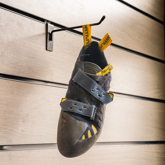 A climbing shoe, La Sportiva Tarantula Boulder, hanging on a black hook against a textured metallic background.