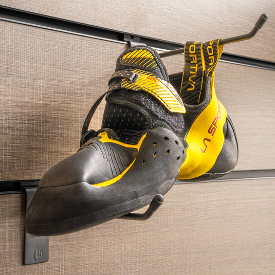 A climbing shoe, La sportiva Solution comp on a black hook and with toe box balanced on another black hook against a textured metallic background.