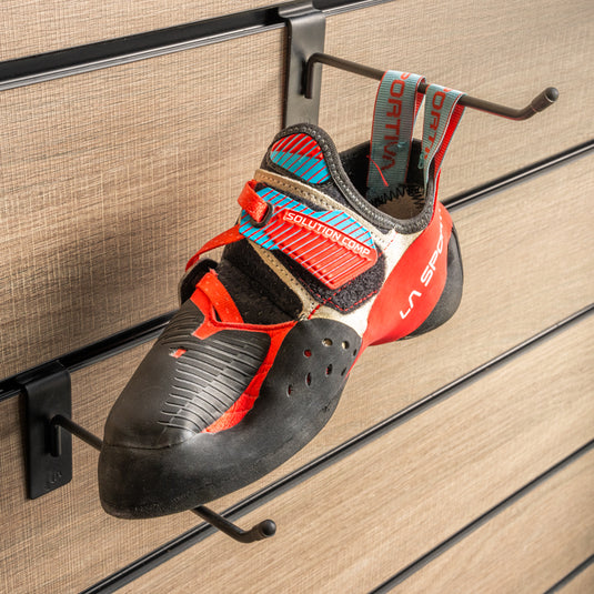 A climbing shoe, La sportiva Solution comp womens on a black hook and with toe box balanced on another black hook against a textured metallic background.