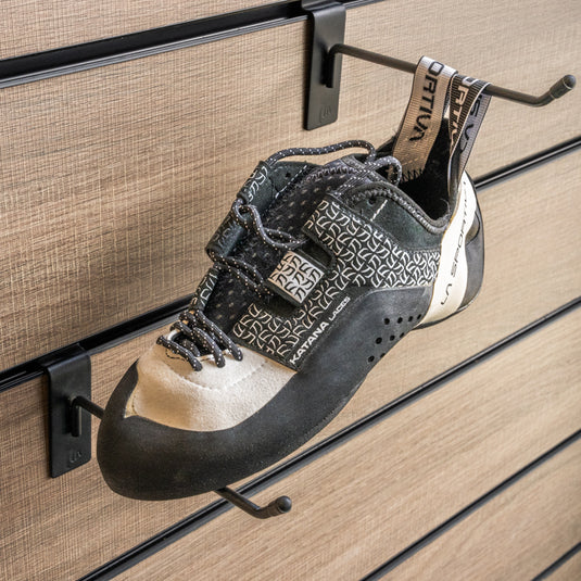 A climbing shoe, La sportiva Katana lace women hanging on a black hook and with toe box balanced on another black hook against a textured metallic background.