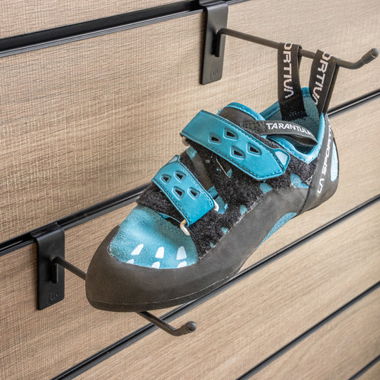 A climbing shoe, La sportiva Tarantula Women on a black hook and with toe box balanced on another black hook against a textured metallic background.
