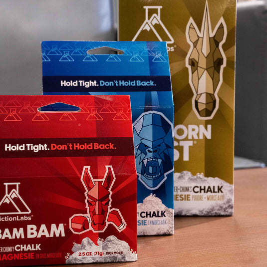 Three packages of FrictionLabs chalk products in red, blue, and gold stand on a wooden surface, displaying a horse, a gorilla and a unicorn geometric animal illustrations.