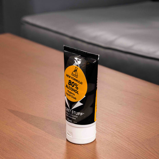 One 75ml tube of Friction Labs Secret Stuff Liquid Chalk in black and white design with a circular orange sticker with 80% alcohol text. The tube is displayed on a wooden surface turned to the side.