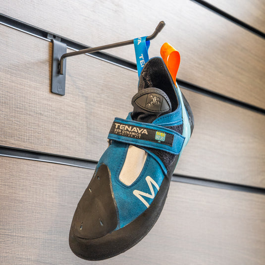 A climbing shoe, Tenaya Mastia hanging on a black hook against a textured metallic background.