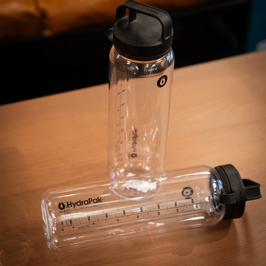 Two Recon water bottles displayed on wooden surface. Both are clear with one stood up and one lay down.