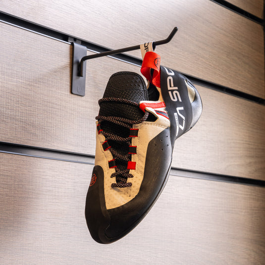 A climbing shoe, La Sportiva Genius, hanging on a black hook against a textured metallic background.