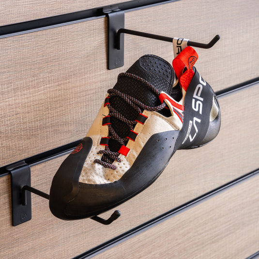 A climbing shoe, La sportiva Genius hanging on a black hook and with toe box balanced on another black hook against a textured metallic background.