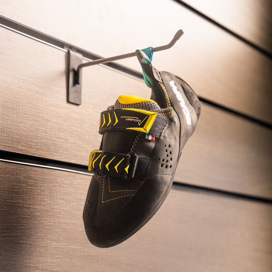 A climbing shoe, Scarpa Vapour V, hanging on a black hook against a textured metallic background.