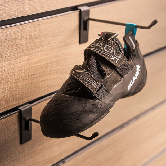 A climbing shoe, Scarpa Drago XT, hanging on a black hook and with toe box balanced on another black hook against a textured metallic background.