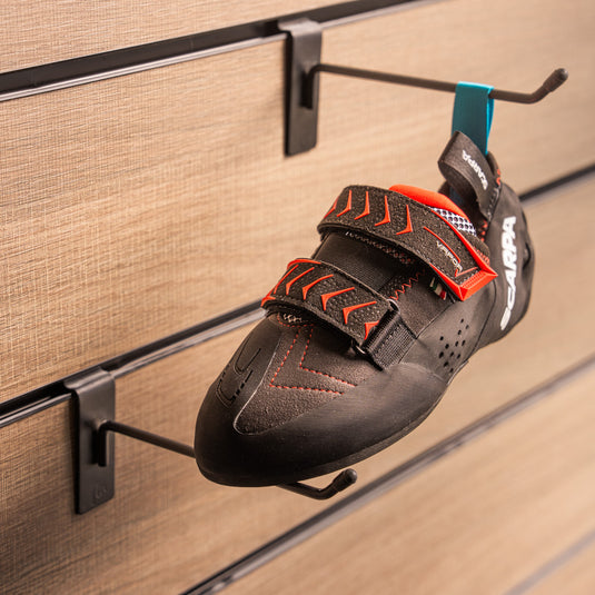 A climbing shoe, Scarpa Vapour V LV hanging on a black hook and with toe box balanced on another black hook against a textured metallic background.