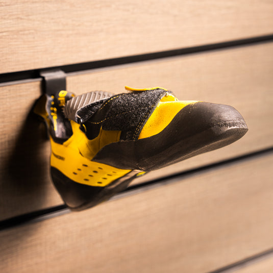A La Sportiva Katana velcro climbing shoe with a yellow and black heel strap loop, hanging on a wall-mounted holder against a textured background.