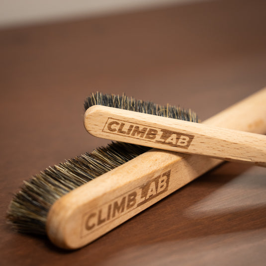 Two wooden climbing brushes with "CLIMBLAB" engraved, resting on a wooden surface, showcasing their bristle details.