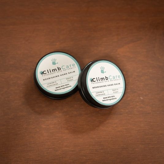 Two containers of ClimbCare nourishing hand balm laid on wooden surface in pale blue and white design.