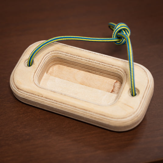 A wooden rectangular offcut handboard with a cut out in the centre. Green and yellow cord threaded through either side, resting on wooden surface