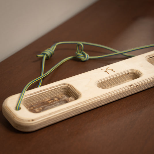 A wooden ash hangboard with various cutouts in the centre with blue and yellow cord threaded through either side. Displayed on wooden surface.