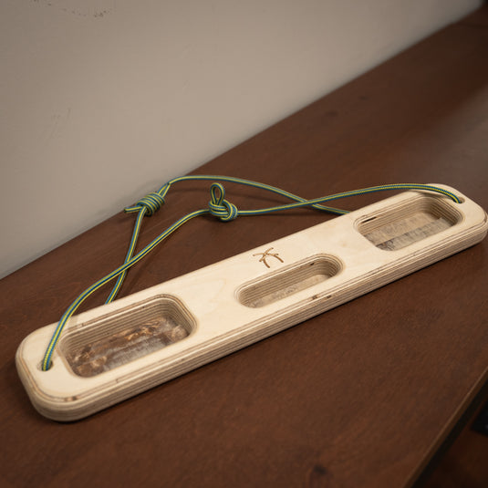 A wooden ash hangboard with various cutouts in the centre with blue and yellow cord threaded through either side. Displayed on wooden surface.