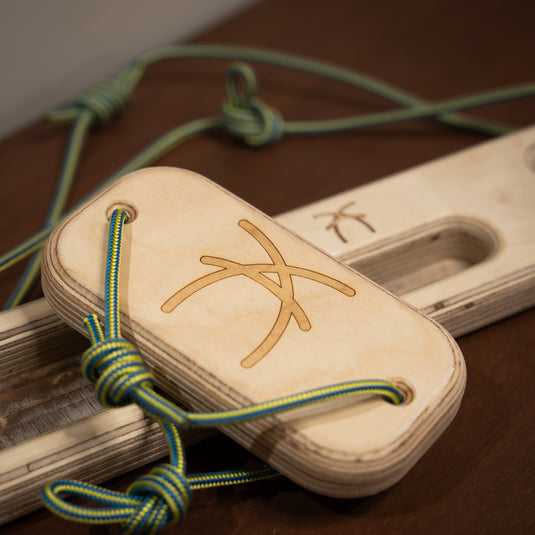 The back of a wooden rectangular offcut hangboard with green and yellow cord threaded through either side, with ash logo on the centre resting on wooden surface