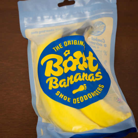 Two yellow Boot bananas attached to blue carabiner inside blue and yellow see-through packaging - laid on wooden surface