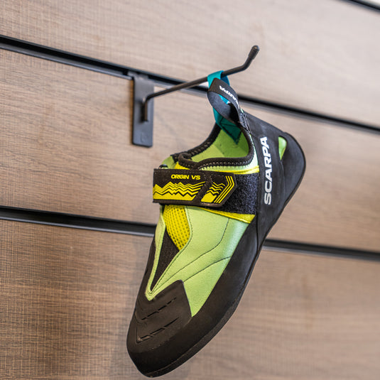 A climbing shoe, Scarpa Origin VS womens, hanging on a black hook against a textured metallic background.