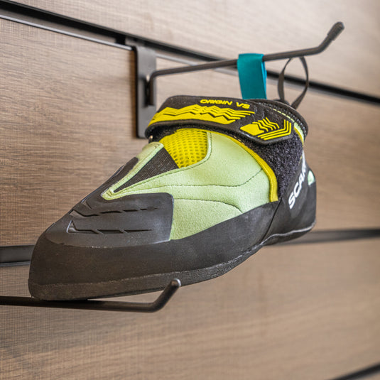 A climbing shoe, Scarpa Origin VS womens, hanging on a black hook and with toe box balanced on another black hook against a textured metallic background.