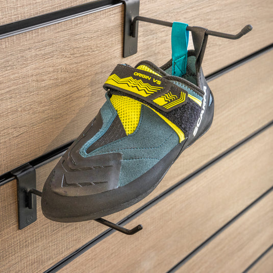 A climbing shoe, Scarpa Origin VS, hanging on a black hook and with toe box balanced on another black hook against a textured metallic background.
