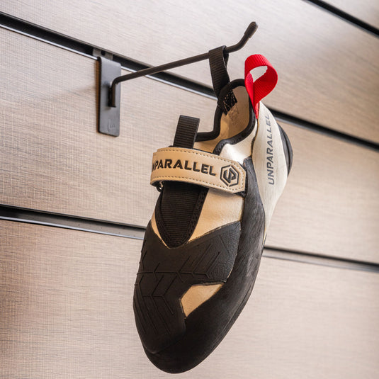 A climbing shoe, Unparallel Qubit hanging on a black hook against a textured metallic background.