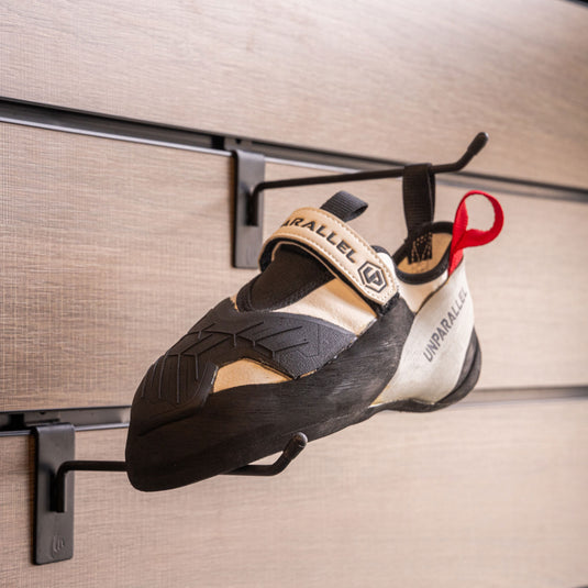 A climbing shoe, Unparallel Qubit hanging on a black hook and with toe box balanced on another black hook against a textured metallic background.