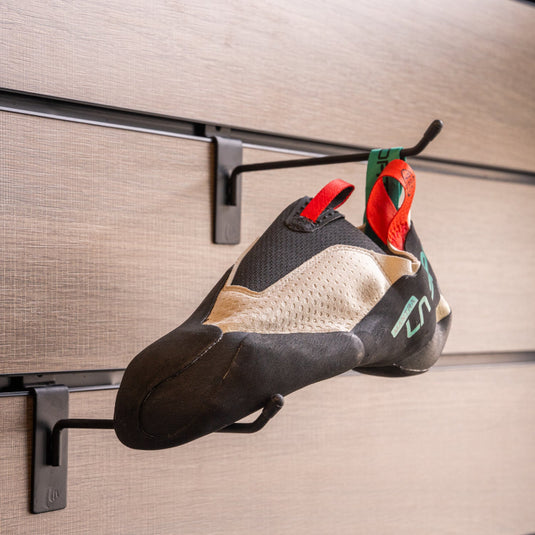 A climbing shoe, La sportiva Mantra hanging on a black hook and with toe box balanced on another black hook against a textured metallic background.