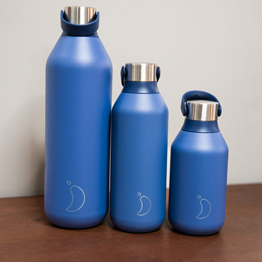 3 blue chillys bottles in different sizes displayed on wooden surface. 1L, 500ml and 350ml all with stainless steel lid.