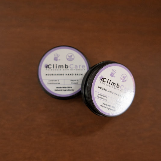 Two containers of ClimbCare nourishing hand balm laid on wooden surface in purple and white design.
